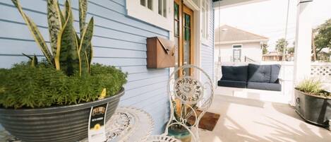 A covered patio with a blue house, a white metal table, a metal chair, a hanging lantern, and a mailbox.