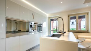 Cocina moderna con muebles blancos, electrodomésticos de acero inoxidable, una isla central con lavamanos y vistas a un edificio de ladrillo a través de amplias ventanas.
