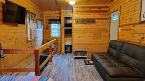 Een knus blokhutinterieur met een houten bank, een zwarte leren bank, een aan de muur bevestigde televisie en een raam met uitzicht op bomen.