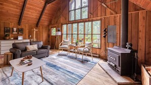 A cozy cabin interior with a wooden ceiling, large windows, a fireplace, a sofa, a coffee table, and a dining area. - Charming A-Frame @ Mt. Rainier~Nature~Relax! Pet Friendly! (Packwood)