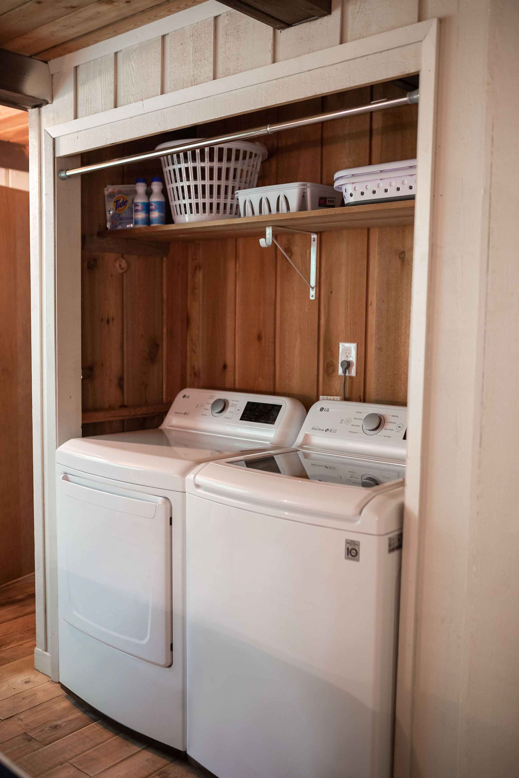A laundry room with two white front-loading washing machines, a white basket, and a shelf with bottles and a white appliance.