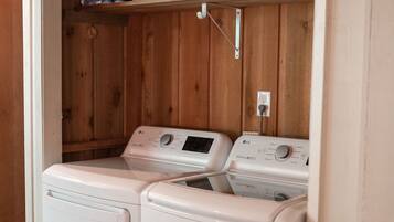 A laundry room with two white front-loading washing machines, a white basket, and a shelf with bottles and a white appliance.