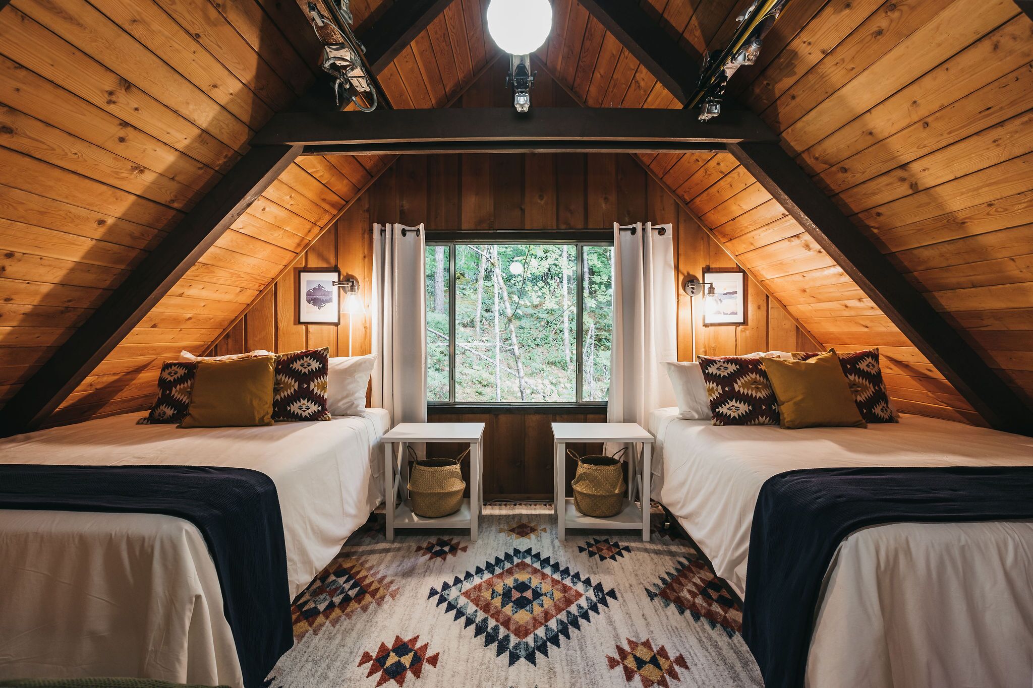 A cozy attic bedroom with two beds, a large window, and a wooden ceiling.
