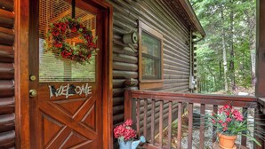 A wooden cabin with a wooden door, a welcome sign, a window, and a balcony with potted plants.