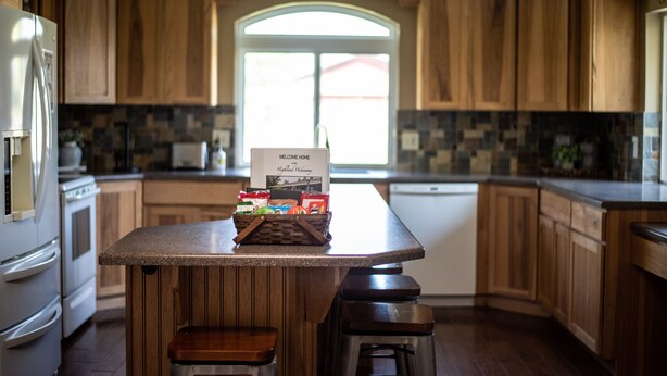 A kitchen with wooden cabinets, a central island with stools, a refrigerator, and a window with a welcome sign. - Highland Hideaway, Room for everyone, central to everything (Highland)