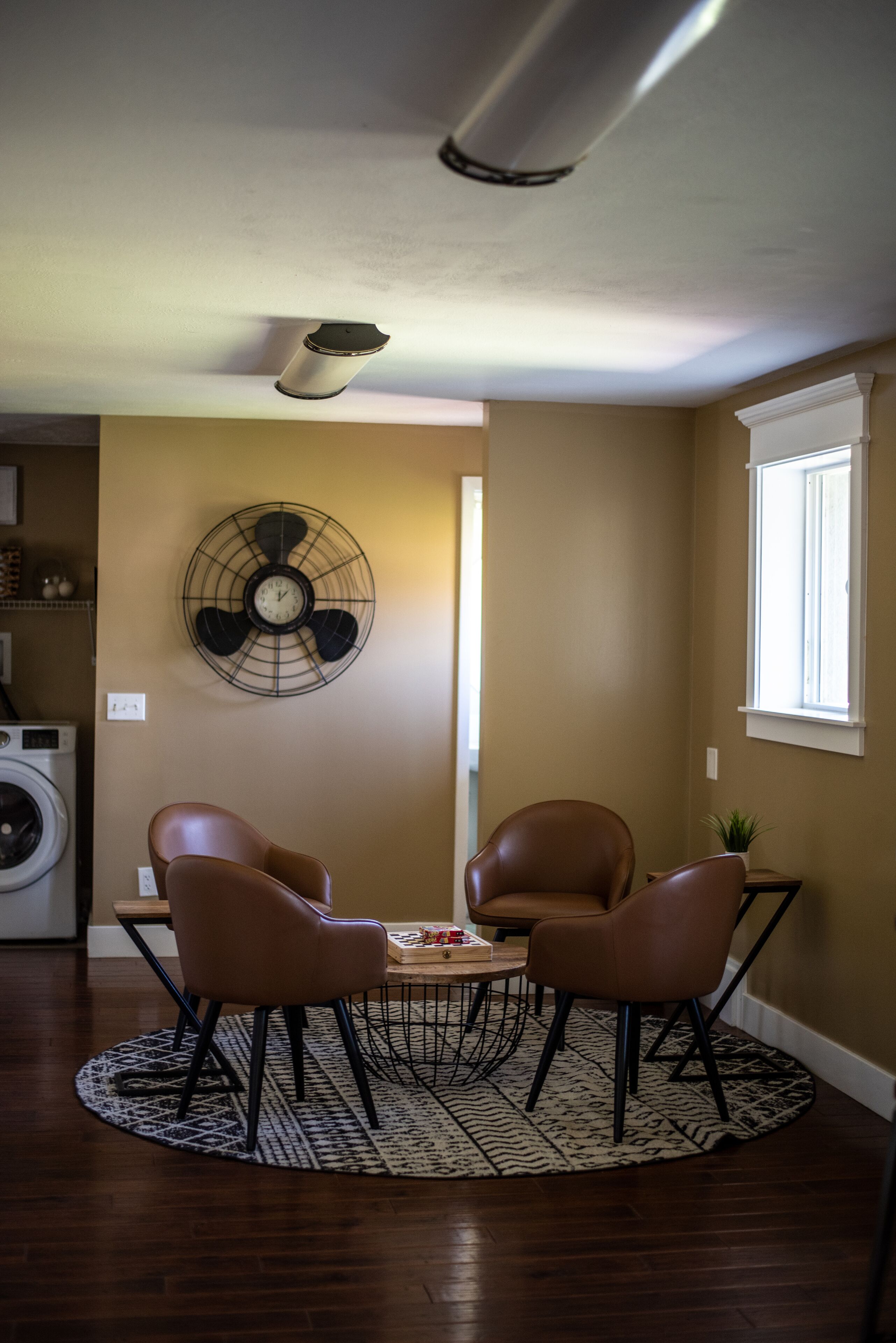 A living room with a round table, two chairs, a fan, and a washing machine in the background.