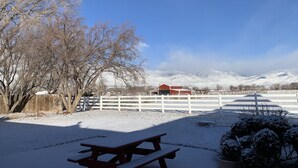 Un área de picnic cubierta de nieve con un banco de madera, una cerca blanca y un edificio rojo al fondo.
