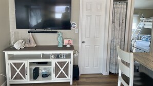 Sala de estar con una televisión de pantalla plana, una mesita consola blanca con un letrero que dice 'BEACH' y una puerta, con una cama literera al fondo.