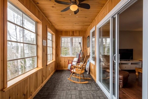 A wooden interior with large windows, a ceiling fan, and a rocking chair. - Modern 4BR Lakefront Cottage w/ Hot Tub & Dock (Emsdale)