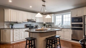 A modern kitchen with white cabinets, a central island with stools, a stainless steel range, and a window providing natural light. - Modern 4BR Lakefront Cottage w/ Hot Tub & Dock (Emsdale)