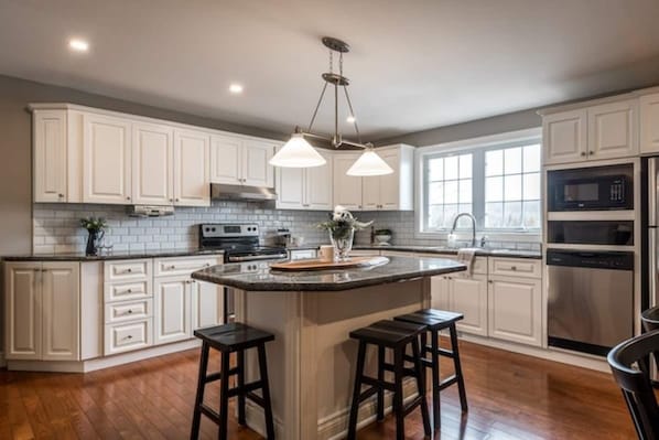 A modern kitchen with white cabinets, a central island with stools, a stainless steel range, and a window providing natural light. - Modern 4BR Lakefront Cottage w/ Hot Tub & Dock (Emsdale)