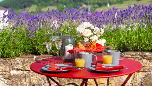 Una tavola rossa apparecchiata per la colazione, con croissant, un vaso di fiori e sullo sfondo un panorama di campi di lavanda e colline.