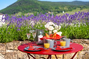 Una mesa roja dispuesta para el desayuno, con croissants, un florero y una vista panorámica de campos de lavanda y colinas al fondo.