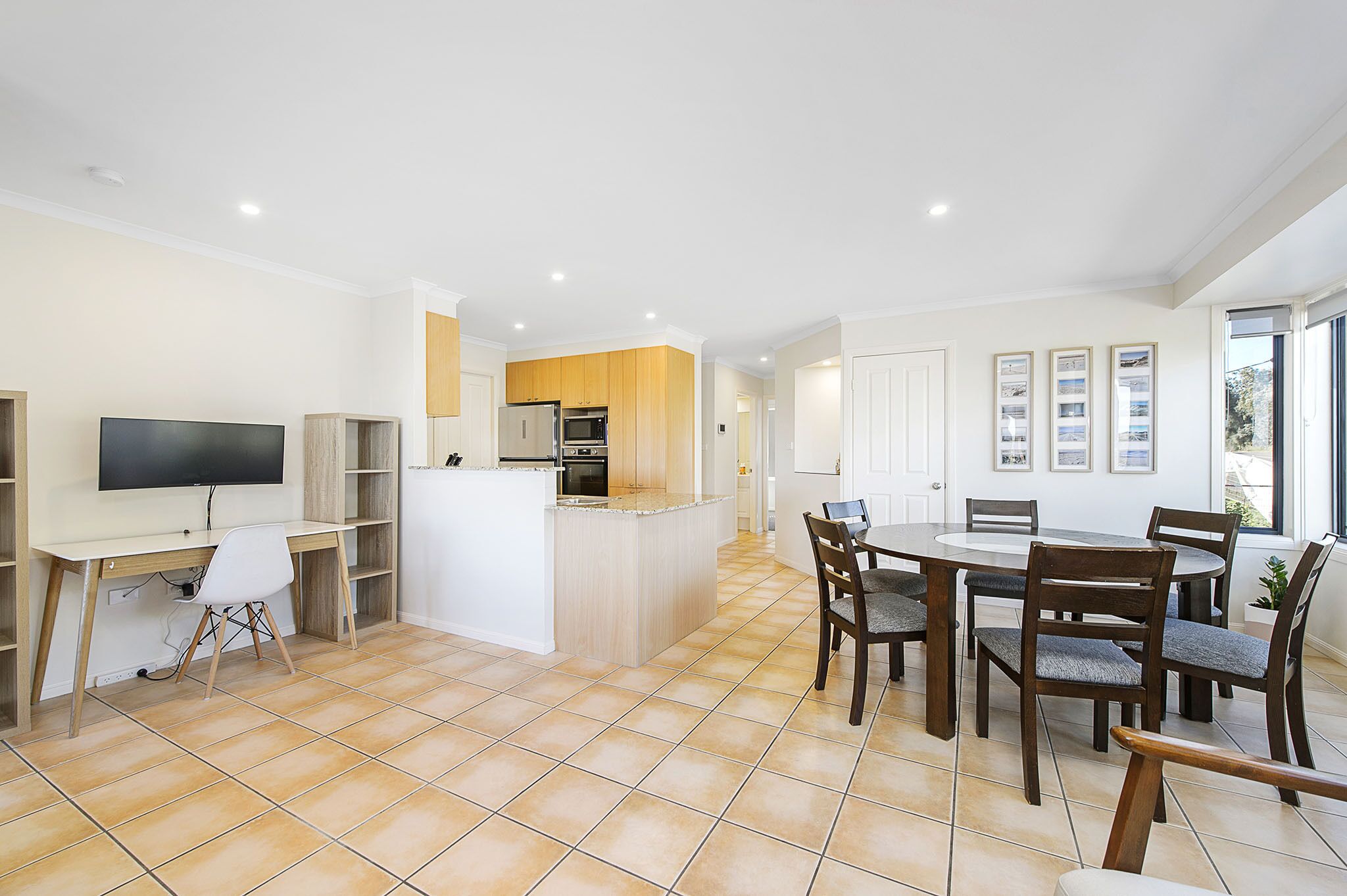 Una cocina moderna con una isla central, un comedor con una mesa redonda y sillas, un televisor montado en un mueble blanco y un piso de baldosas.
