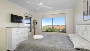 A bedroom with a bed, a TV, a ceiling fan, a dresser, and a window with a view of buildings and trees. - Flynn's Beach Escape ! (Port Macquarie)