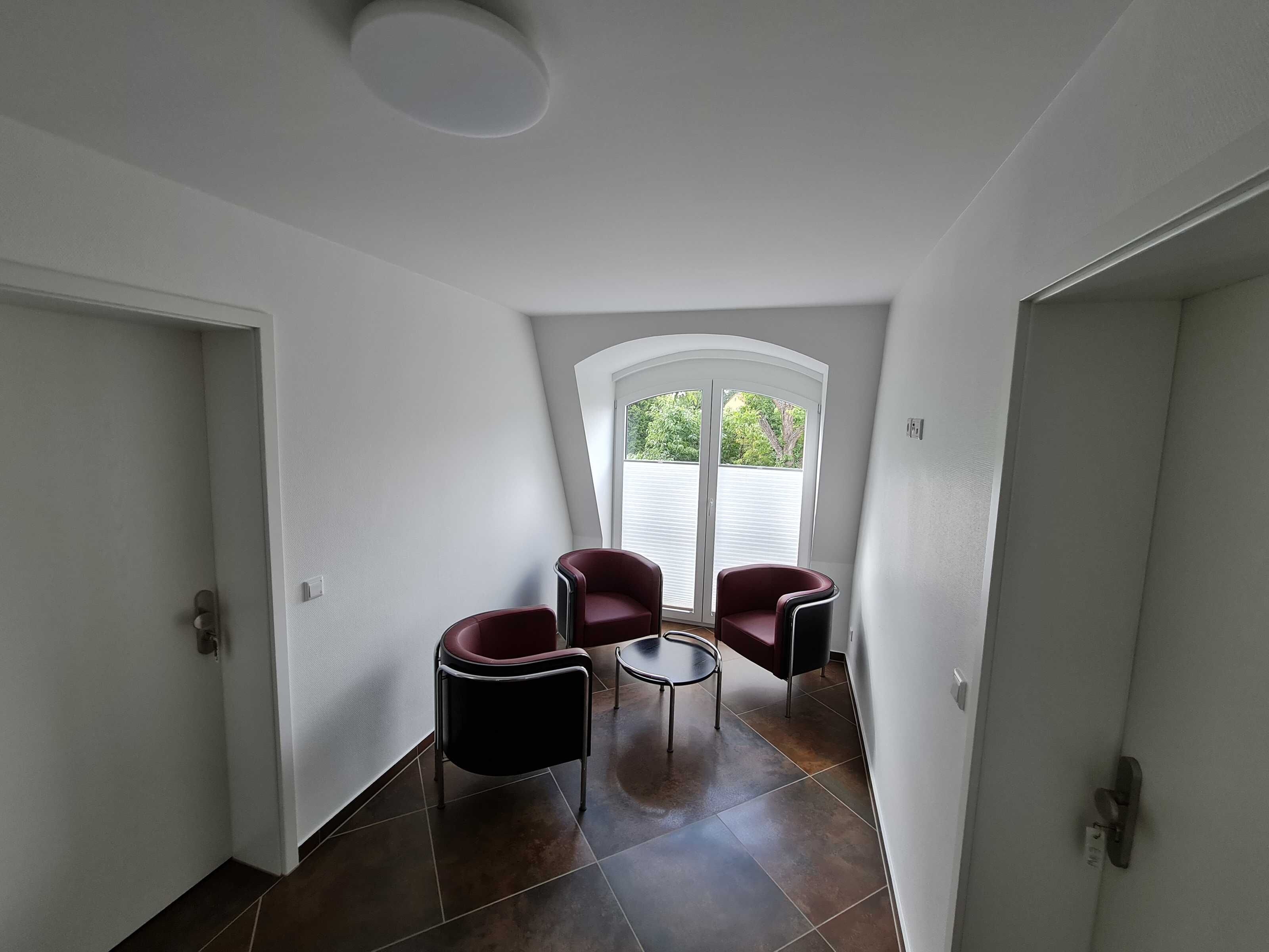A corner room with two red chairs, a small table, and a large window with blinds.