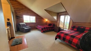 A cozy attic bedroom with a sloped ceiling, two beds with red and black checkered bedding, a gray chair, and a standing fan.
