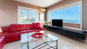 Ein Wohnzimmer mit einem roten Ledersofa, einem Glastisch, einem Flachbildfernseher und einem großen Fenster mit Blick auf das Meer.