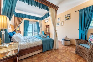 A bedroom with a four-poster bed, a wicker chair, and a bedside table with a lamp.