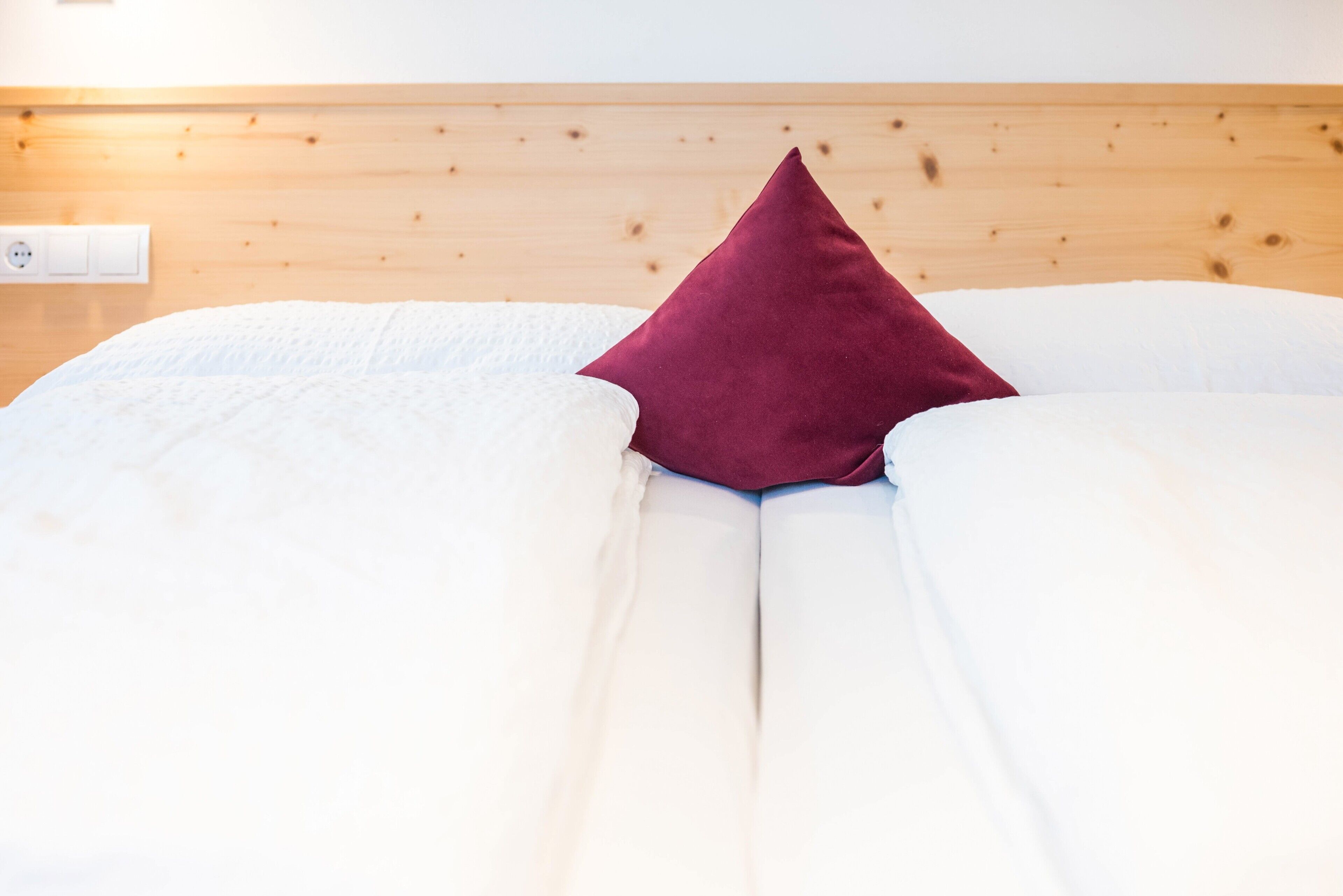 Un lit recouvert d’une couverture blanche, avec un seul oreiller rouge posé contre une tête de lit en bois.