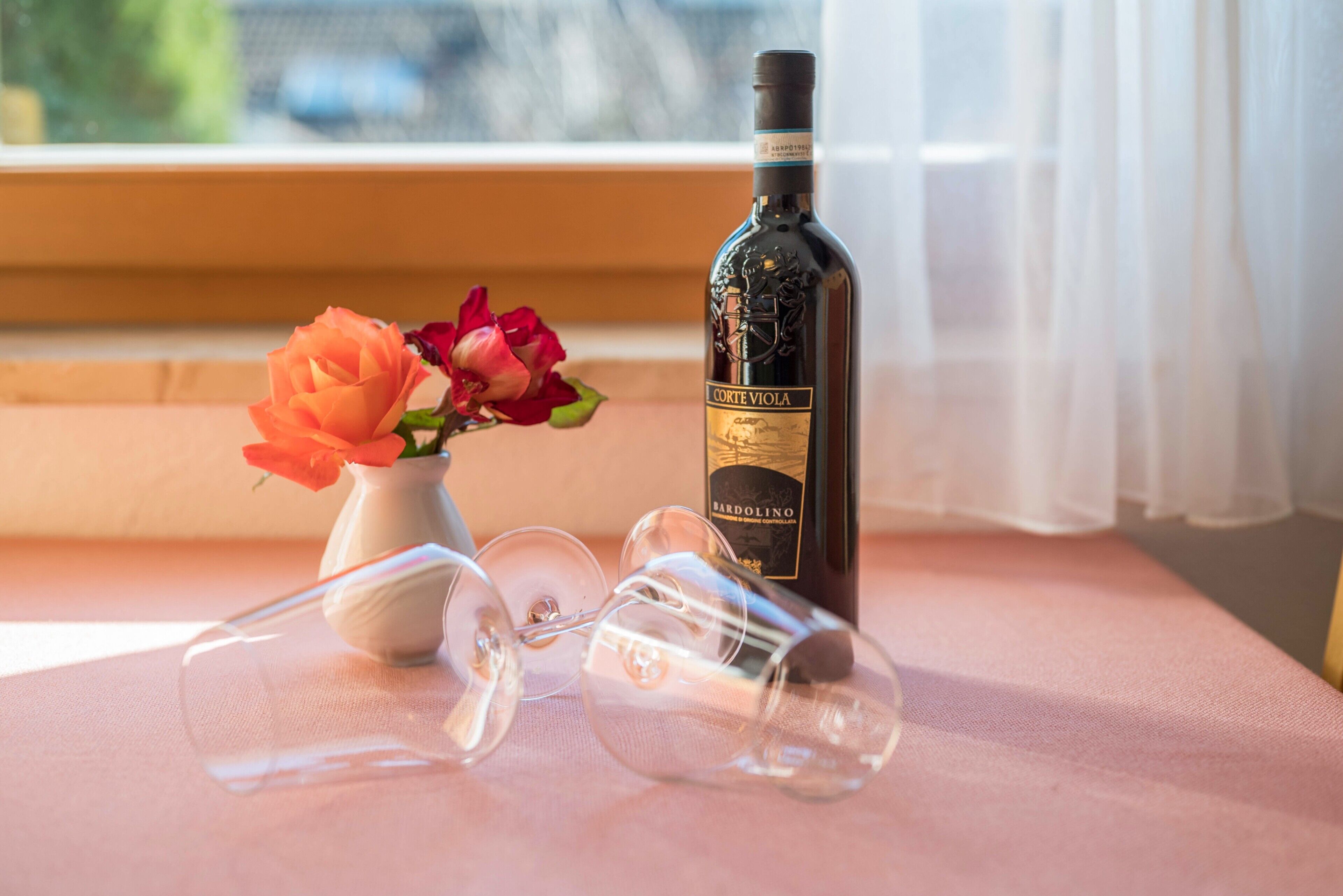 A bottle of wine, a vase with flowers, and wine glasses on a table by a window.