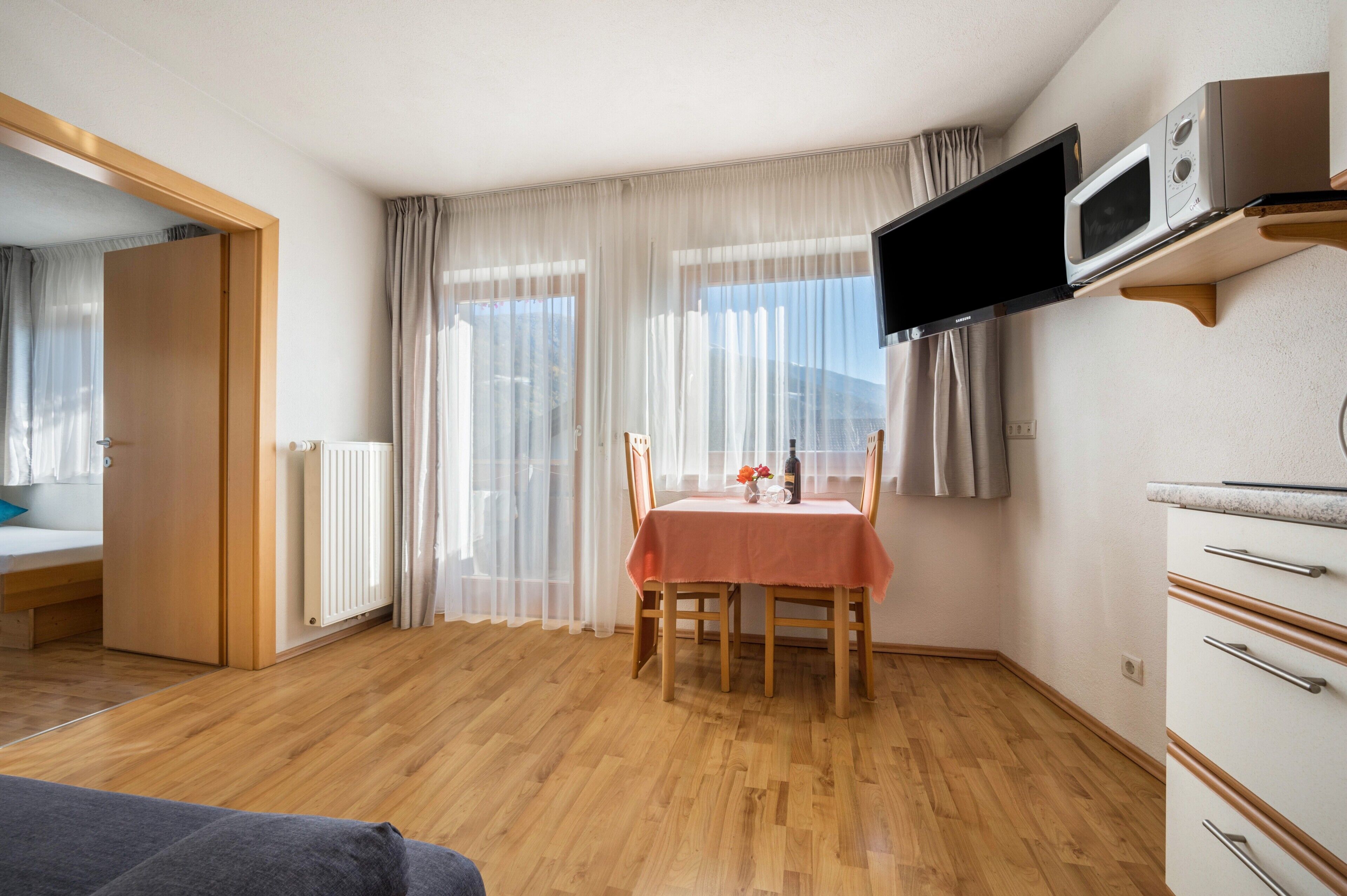 A room with a dining table, chairs, a TV mounted on the wall, a wooden cabinet, and a window with curtains.