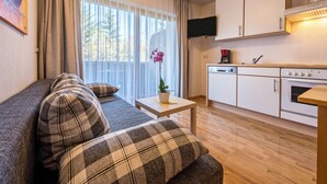 Ein Wohnzimmer mit einer Couch, einem kleinen Tisch mit einer Pflanze, einer Küchenzeile mit Schränken und einem Herd sowie einem an der Wand befestigten Fernseher.