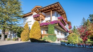 Un bâtiment à deux étages, doté d’un toit en pente, de balcons en bois et d’un jardin bien entretenu où poussent diverses espèces d’arbustes et de fleurs.