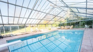 Une piscine intérieure dotée d’un toit en verre, entourée de chaises longues et offrant une vue sur l’extérieur.