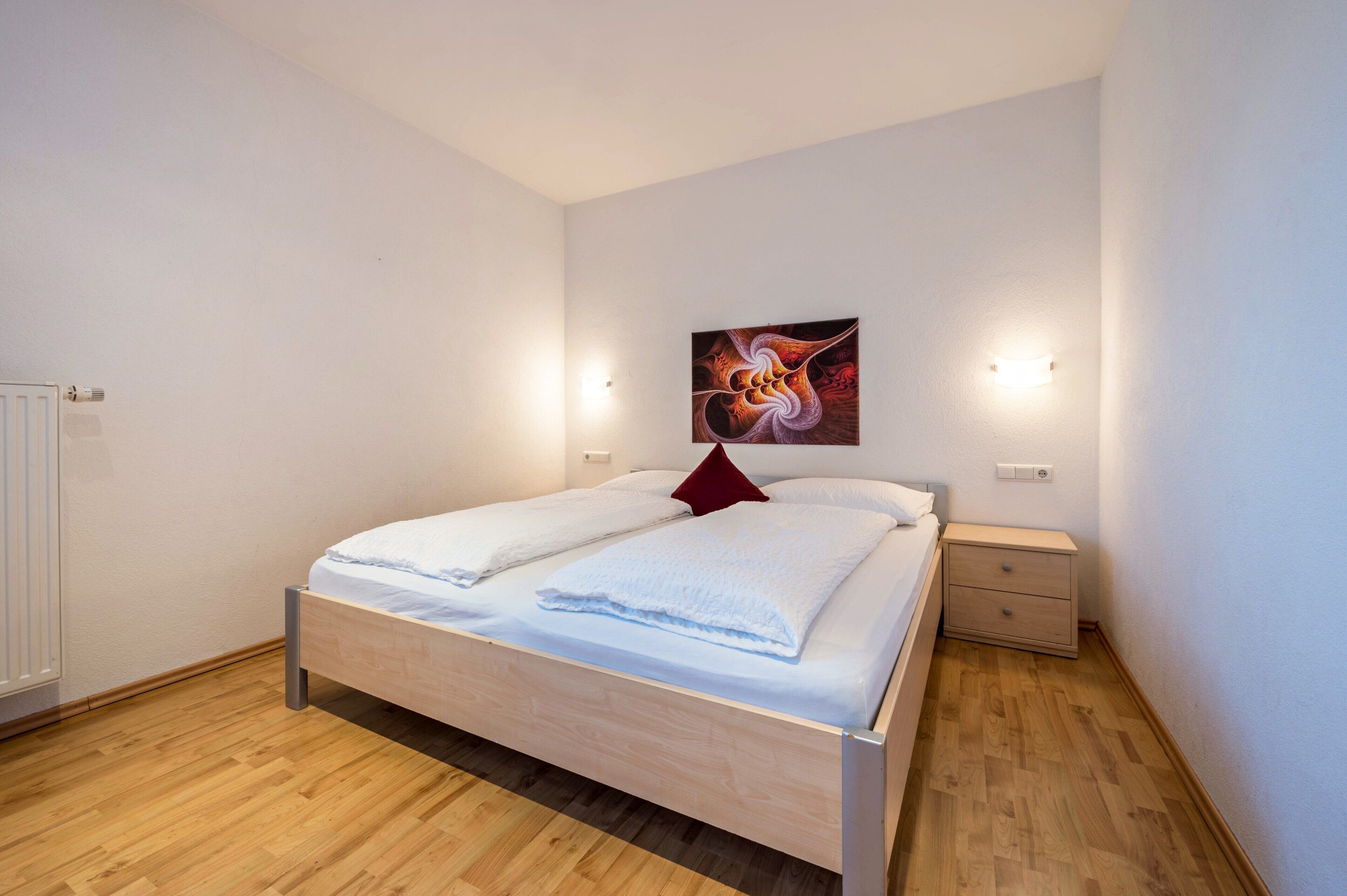 A bedroom with a wooden bed, a red pillow, a nightstand, and a wall-mounted light fixture.