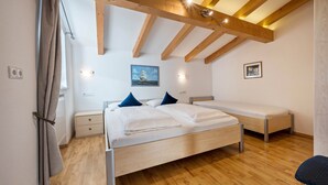 Une chambre avec un plafond en bois, un lit avec une literie blanche, une table de chevet, une commode, une chaise et une fenêtre avec des rideaux.