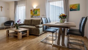 Una sala de estar con un sofá, una mesa de centro, una mesa de comedor con sillas, una silla negra y una cruz en la pared.