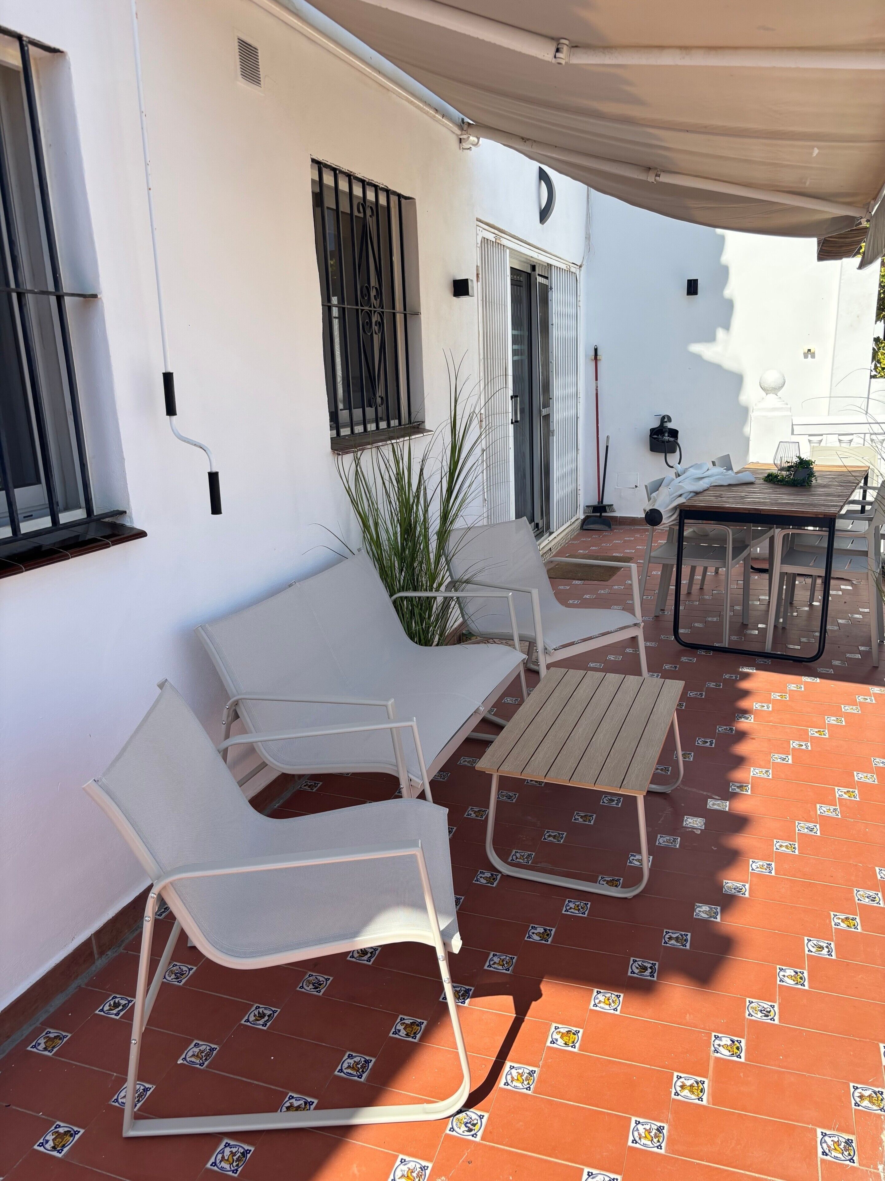 Une terrasse avec des meubles blancs, un sol carrelé et un bâtiment blanc doté d’une fenêtre et d’une porte.