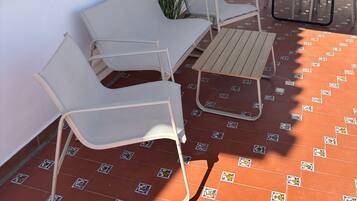 Une terrasse avec des meubles blancs, un sol carrelé et un bâtiment blanc doté d’une fenêtre et d’une porte.