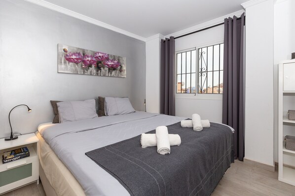 A modern hotel room with a large bed, a bedside table with books, a window with curtains, and a wall art piece. - Apartment "Villa Mar del Plata" with Shared Pool, Wi-Fi, Garden & Terrace (Fuengirola)