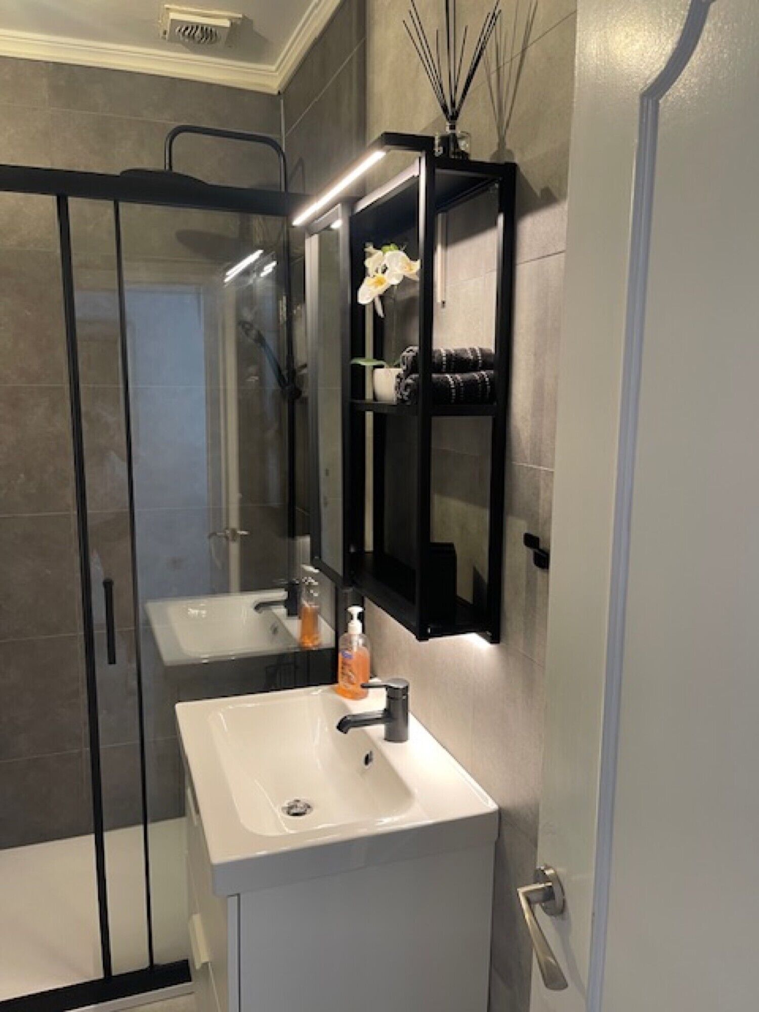 Un baño con cabina de ducha de vidrio, dos lavamanos, un espejo y un estante con artículos de aseo.