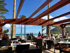 Un espace extérieur aménagé pour les repas, avec des tables et des chaises, une vue sur l’océan et des palmiers.