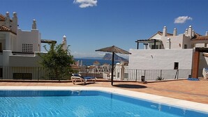 Piscina con sillas de descanso, una sombrilla de paja y vistas a edificios y montañas.