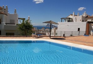 A swimming pool with lounge chairs, a thatched umbrella, and a view of buildings and mountains.