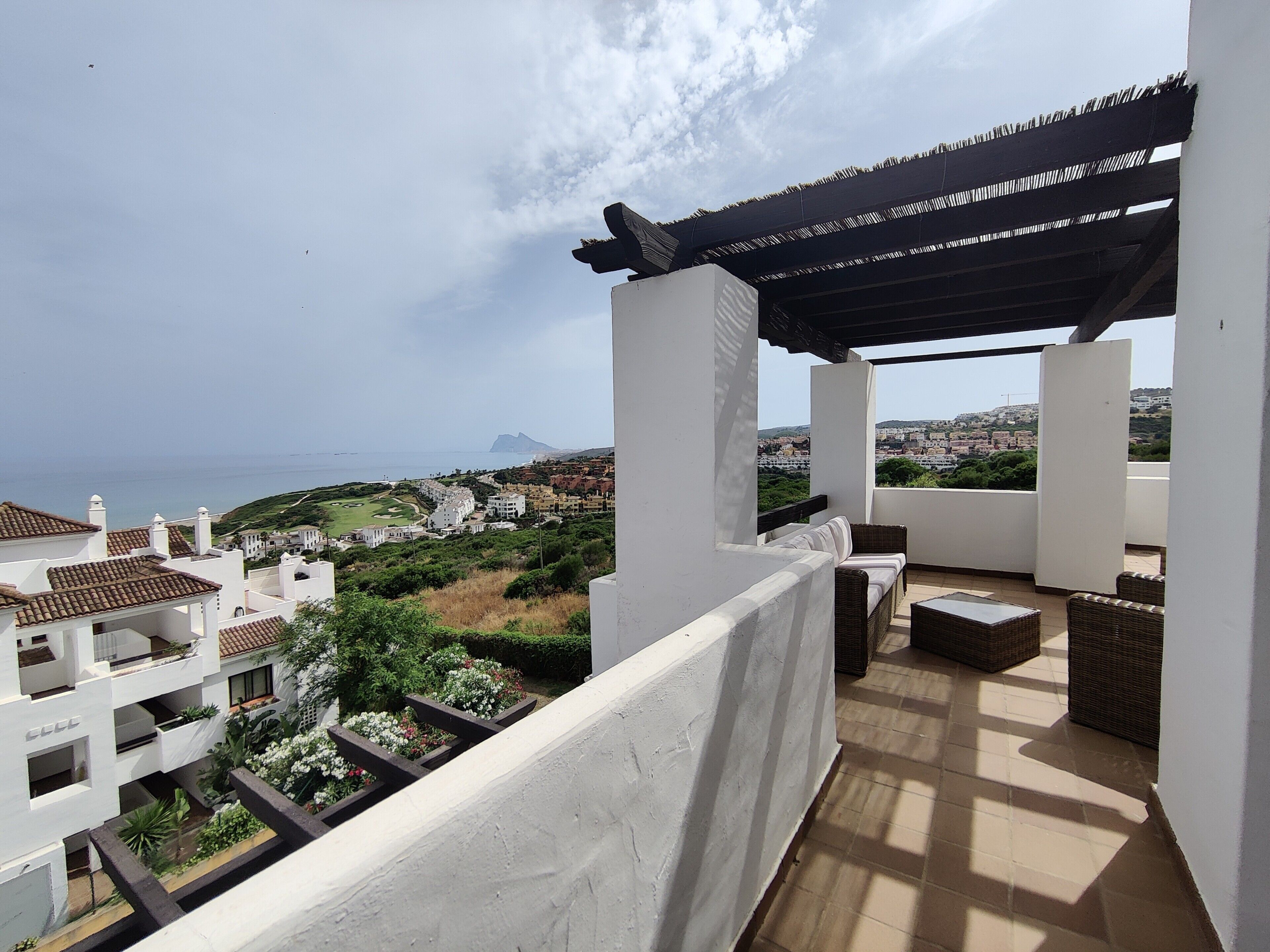 Un balcón con mobiliario exterior, una pérgola y vistas al mar y edificios al fondo.