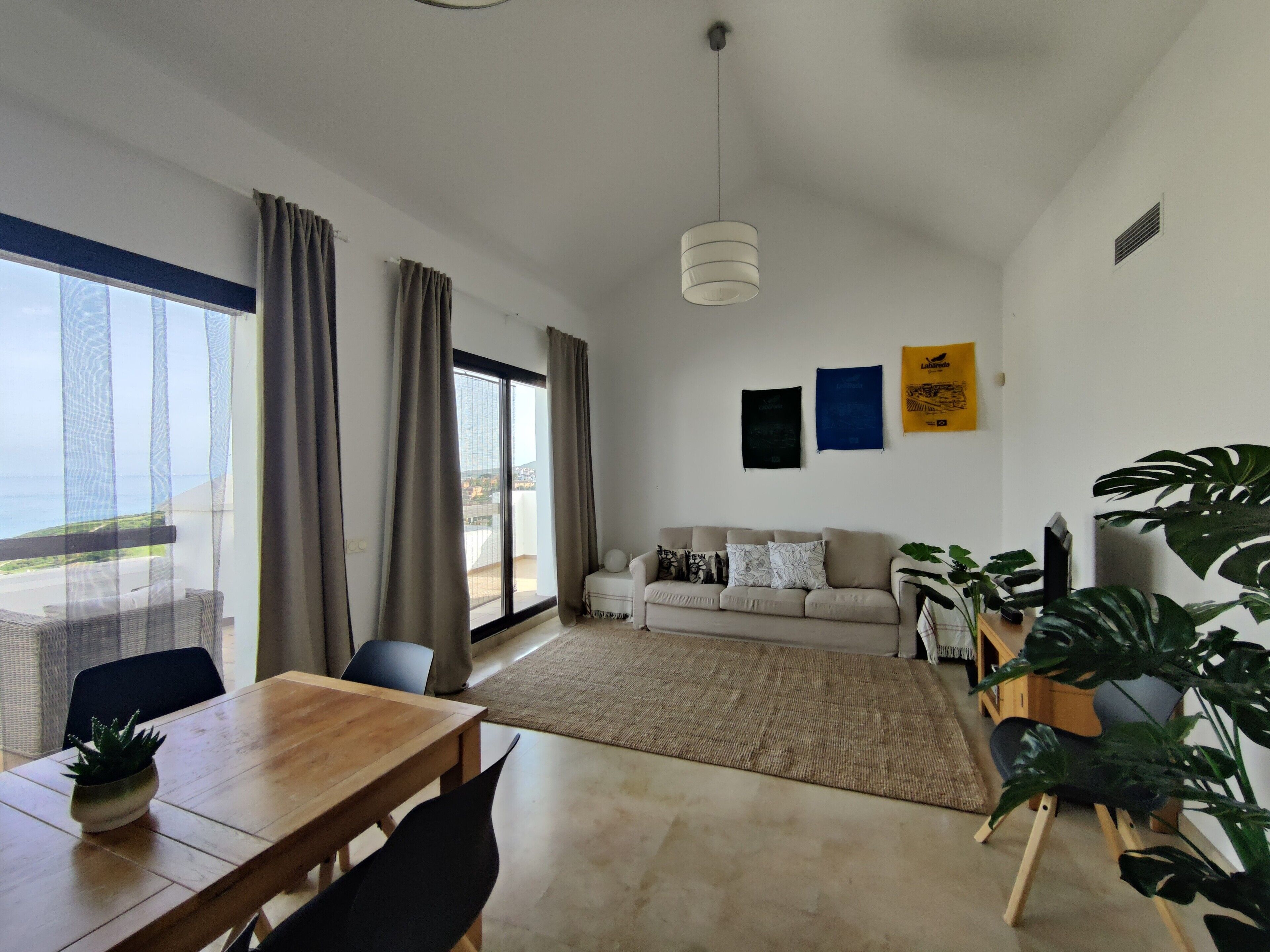 A modern living room with a sofa, dining table, and chairs, a large window with curtains, and a view of the sea.