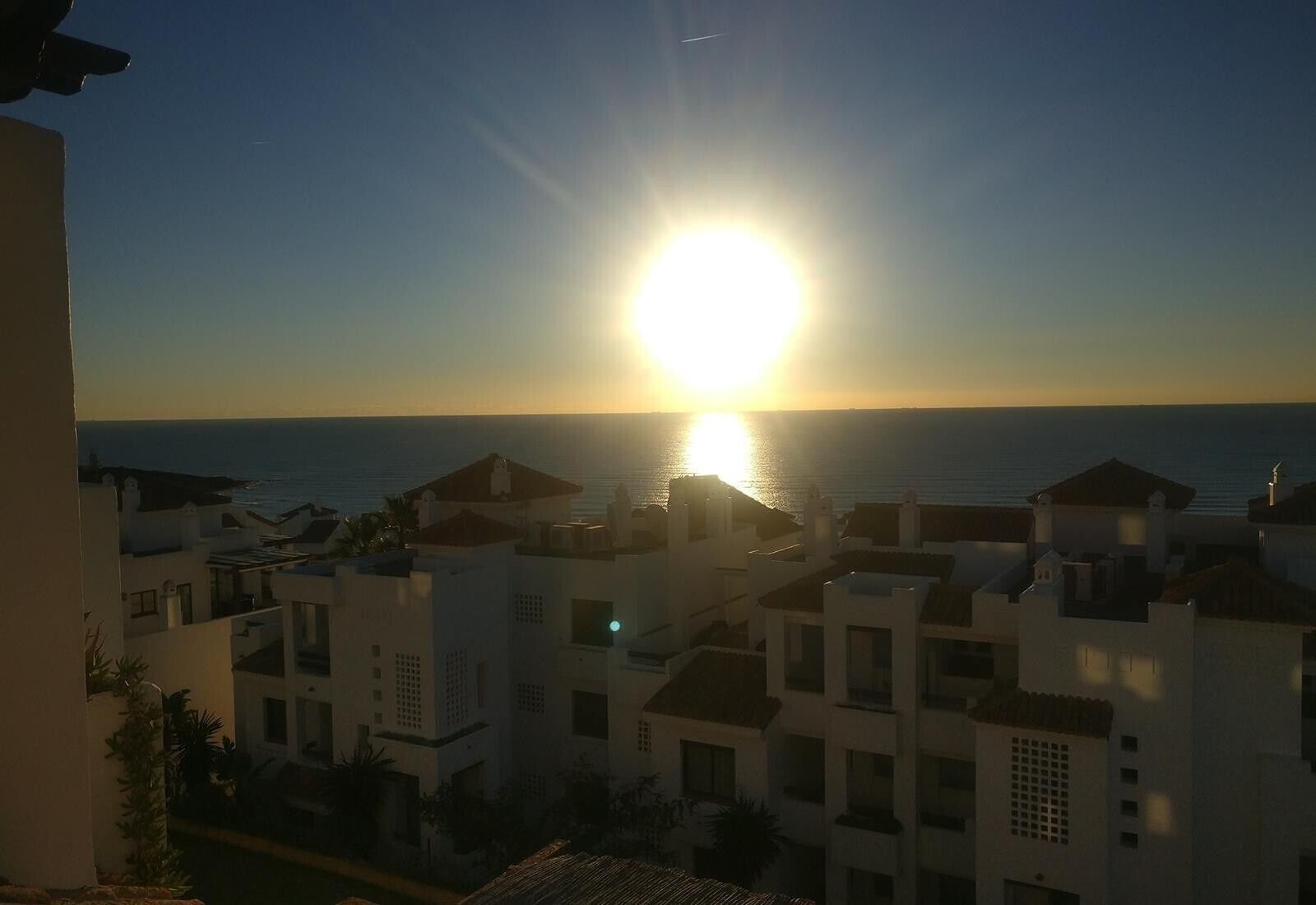 Vista costera con edificios y el sol poniéndose sobre el mar.