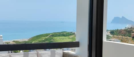 A balcony with a wicker sofa and a glass table, overlooking a coastal landscape with a golf course and buildings.