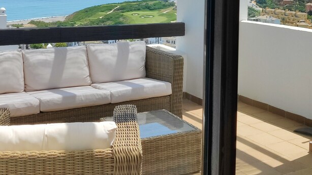 Un balcón con sofá de mimbre y una mesa de vidrio, con vistas a un paisaje costero que incluye un campo de golf y edificios.