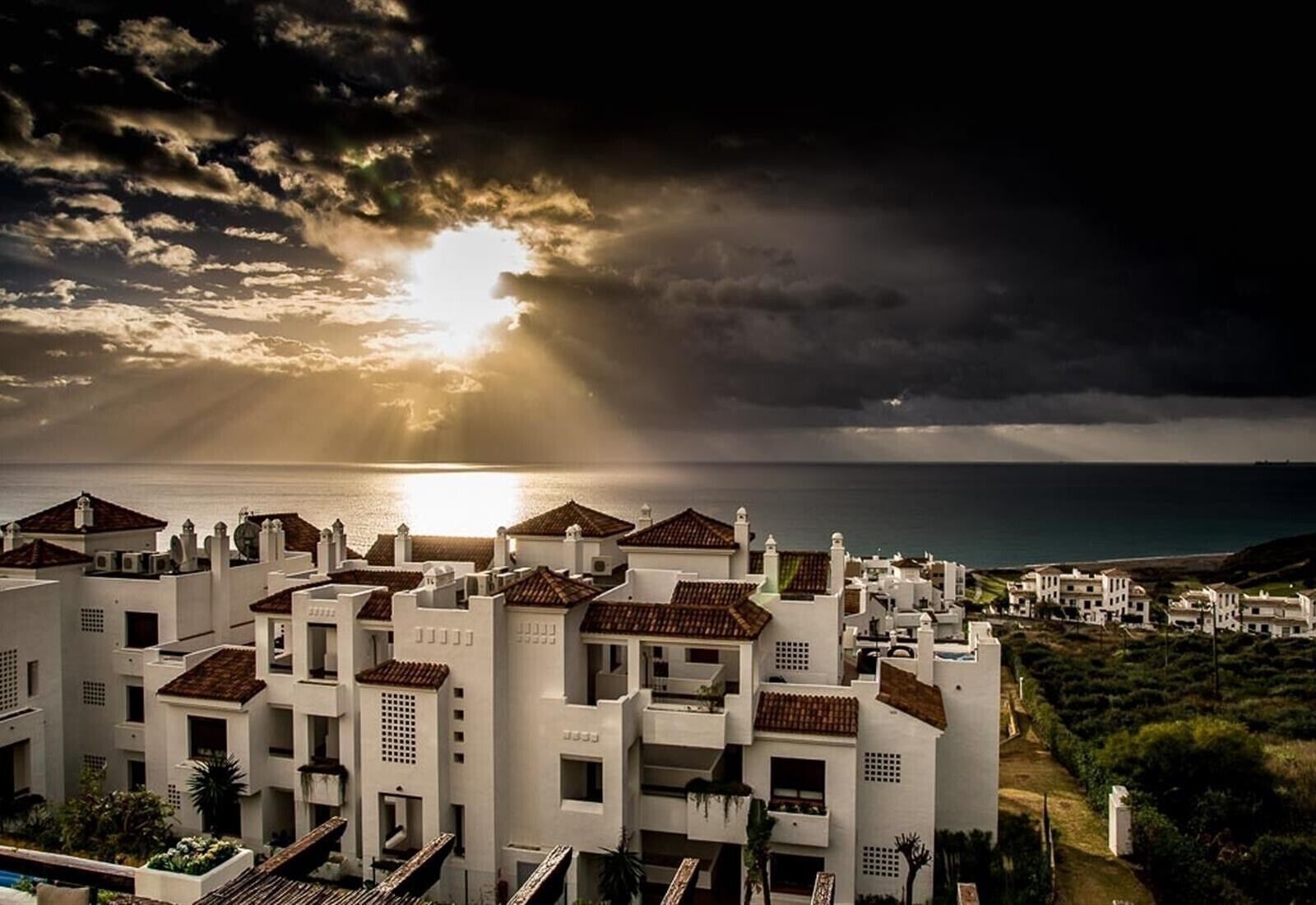 Una zona residencial costera con edificios blancos, algunos con tejados de tejas rojas, y vista al mar.