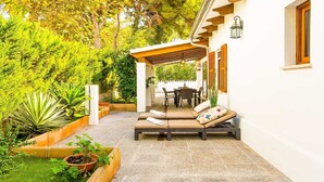 A covered outdoor seating area with lounge chairs and a table, surrounded by greenery and a white building.