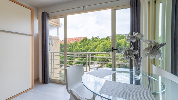 Een moderne kamer met een glazen tafel, witte stoelen, een vaas met bloemen en een balkon met uitzicht op bomen en gebouwen.