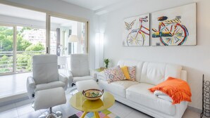 Een moderne woonkamer met een witte bank, twee witte fauteuils, een gele salontafel en een groot raam met uitzicht op groen.