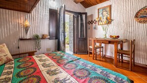 Une pièce avec un plafond en bois, un lit recouvert d’une couverture aux couleurs vives, un bureau avec des chaises et une porte donnant sur l’extérieur.