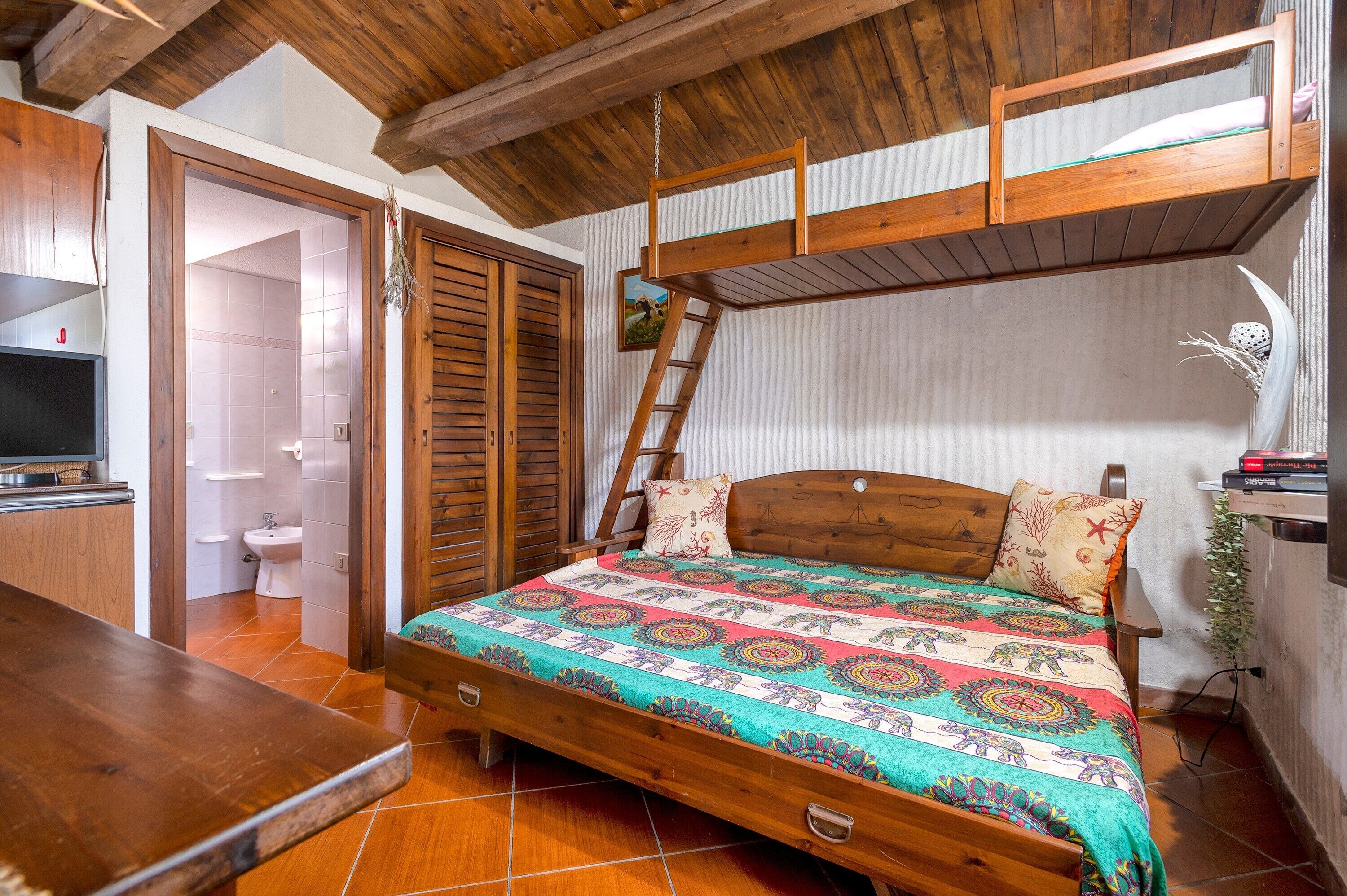 A wooden bunk bed with a colorful quilt, a wooden desk, and a bathroom with a toilet and sink.
